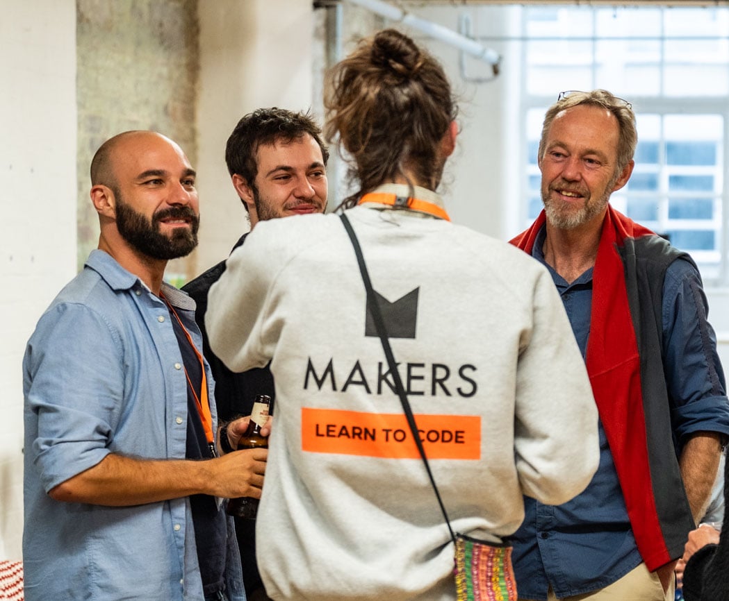 3 men across a range of ages talk with a Makers tutor in branded grey jumper