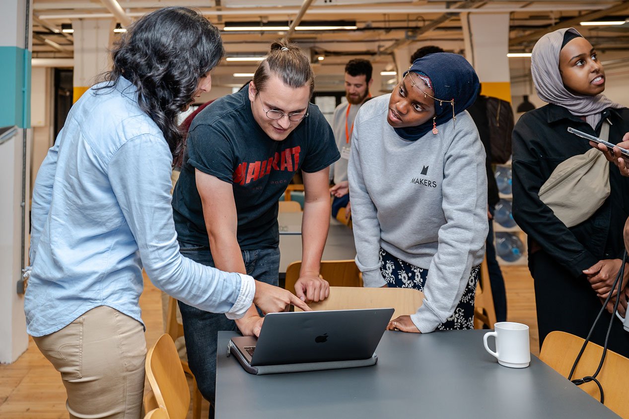 A Makers tutor listens intently to a student while another looks at a laptop. An Asian lady stands to the side in another conversation (small B)