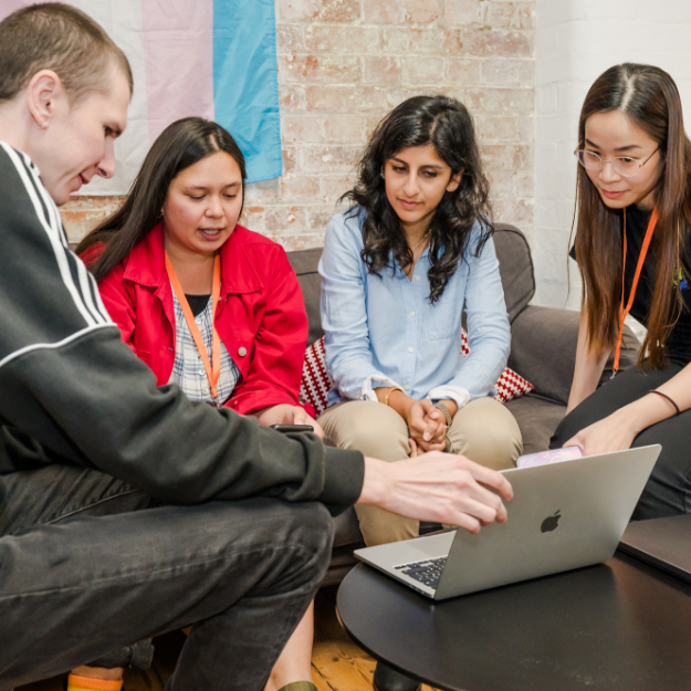 A group of 4 young people talk together around a laptop on comfy chairs.
