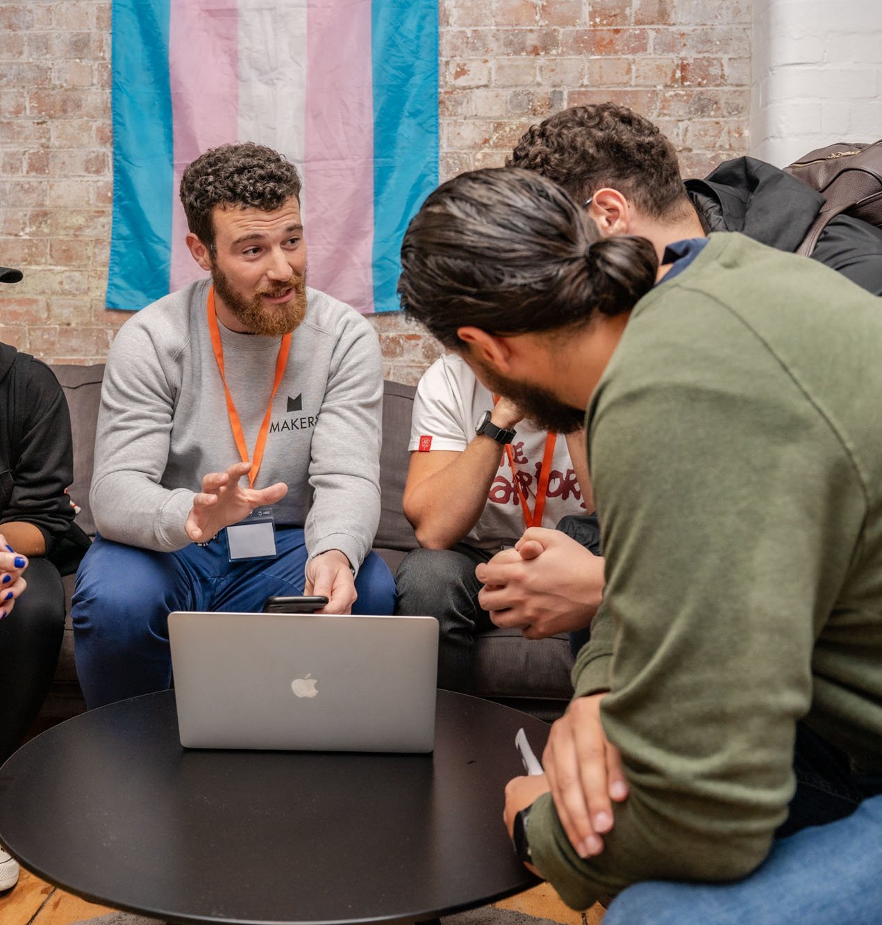 A man with a ponytail looks away from the camera into a huddle of people listening to a tutor with a beard, with a laptop at the centre