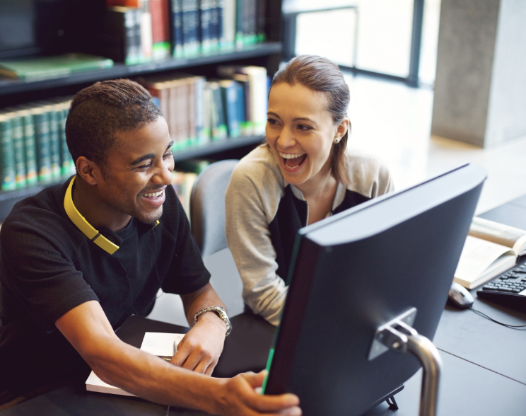 A tutor and student sit laughing at a desk, with long bookshelves behind holding large sets of matching books.