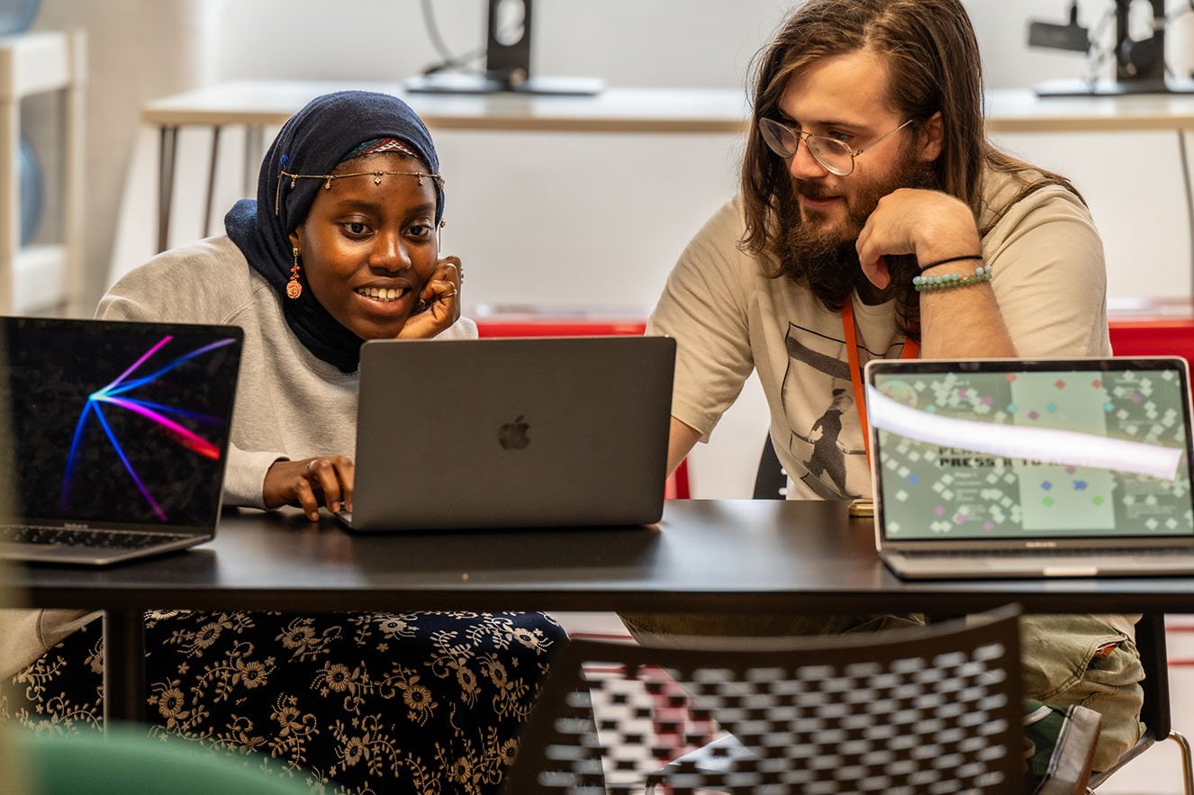 A Makers Tutor is helping a student work on a laptop at a workbench with 2 other laptops open