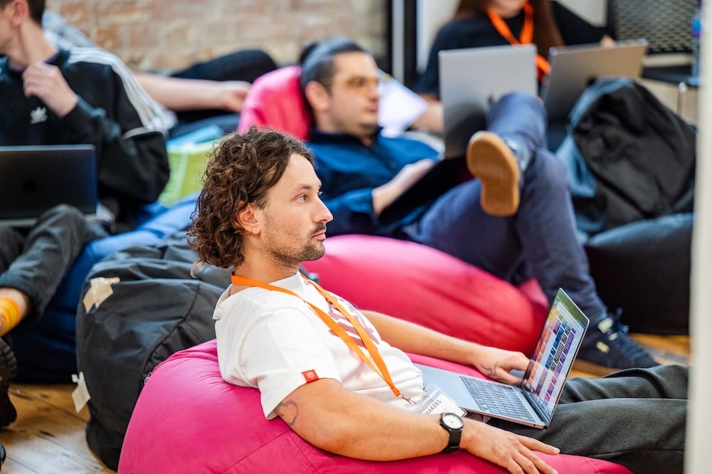 A student sits on a pink beanbag working on a laptop. Other students sit in the background