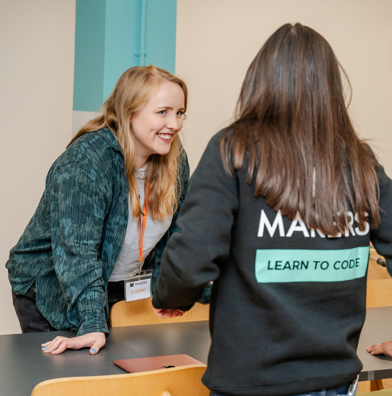 Female Makers tutor with long brown hair over a branded black jumper, talks to a blonde student with a green patterned sweater